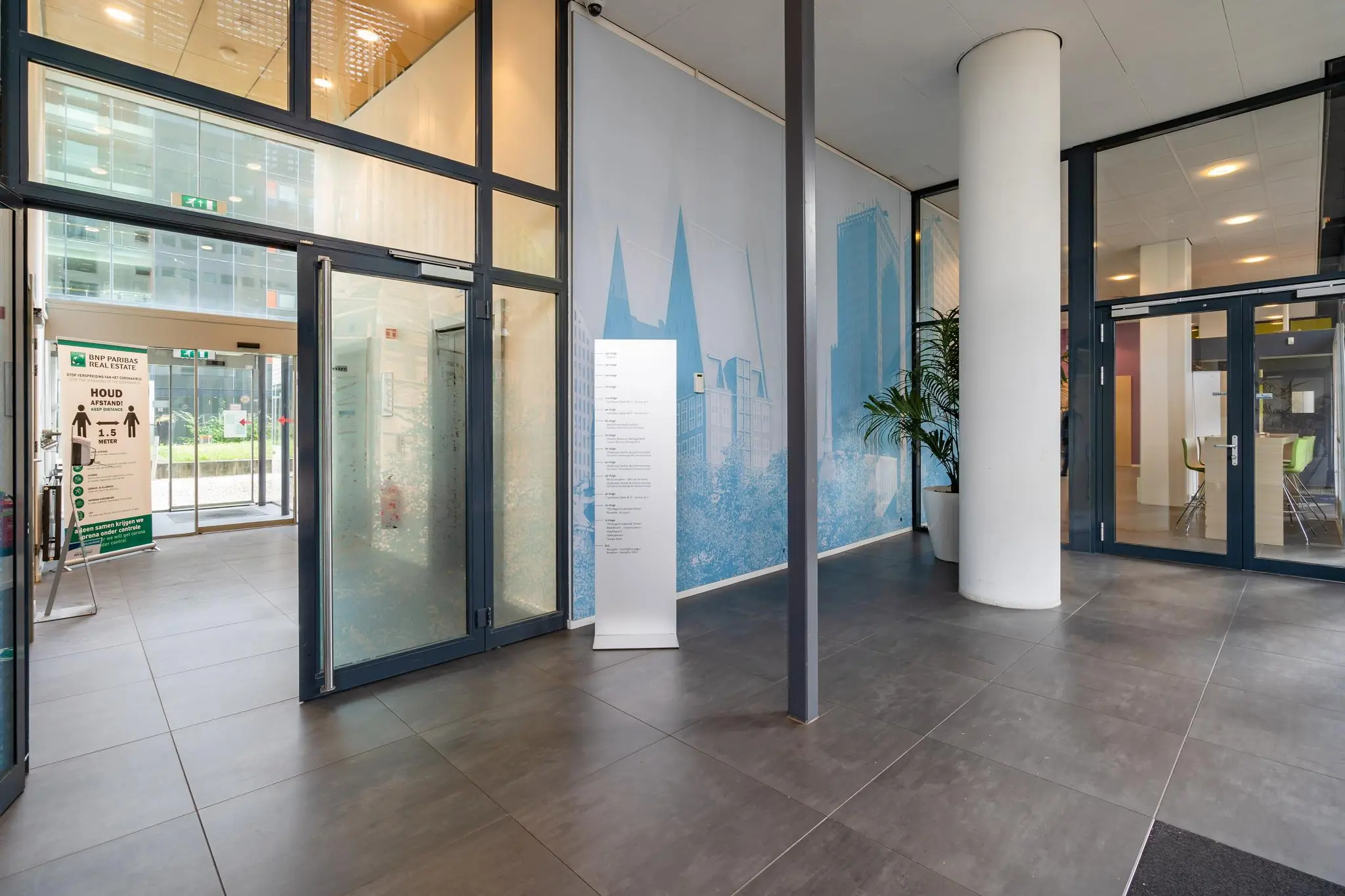 Ingang van een modern kantoorgebouw aan het Johanna Westerdijkplein met glazen deuren, een informatiebord en een coronamaatregelbord.